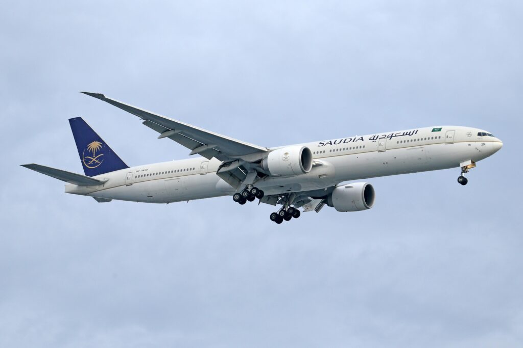 A Saudia Airlines Boeing 777 aircraft flying against a cloudy sky backdrop.
